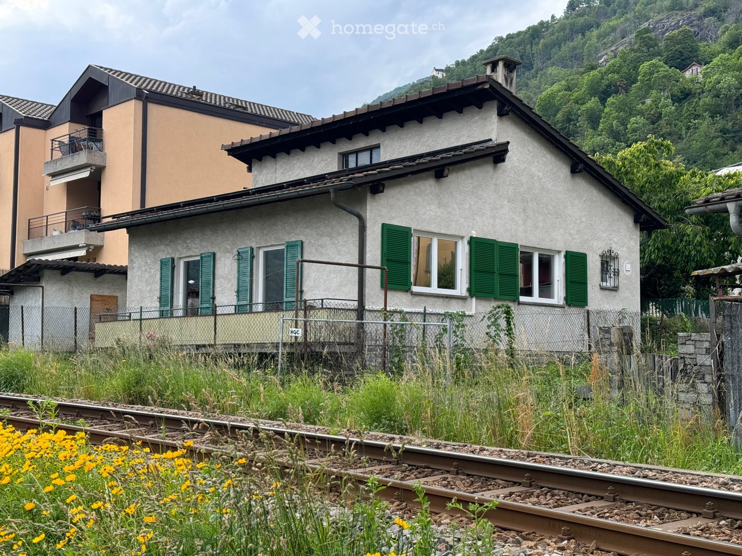 Two-story house with green shutters, white windows, and a balcony overlooking a railway track.