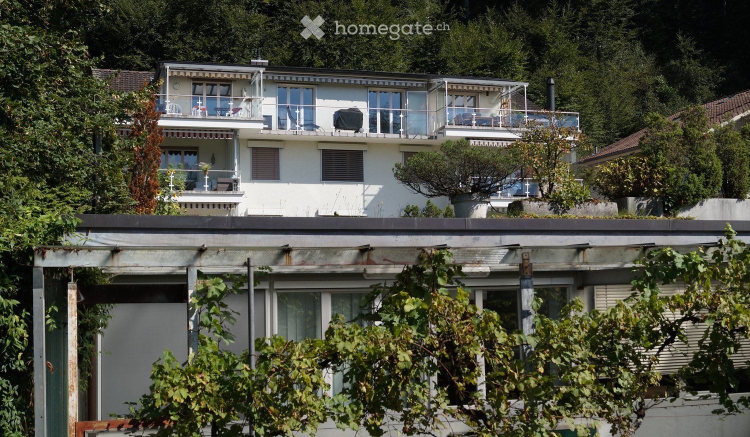 Two-story house with white exterior, multiple balconies, and large windows, surrounded by greenery.