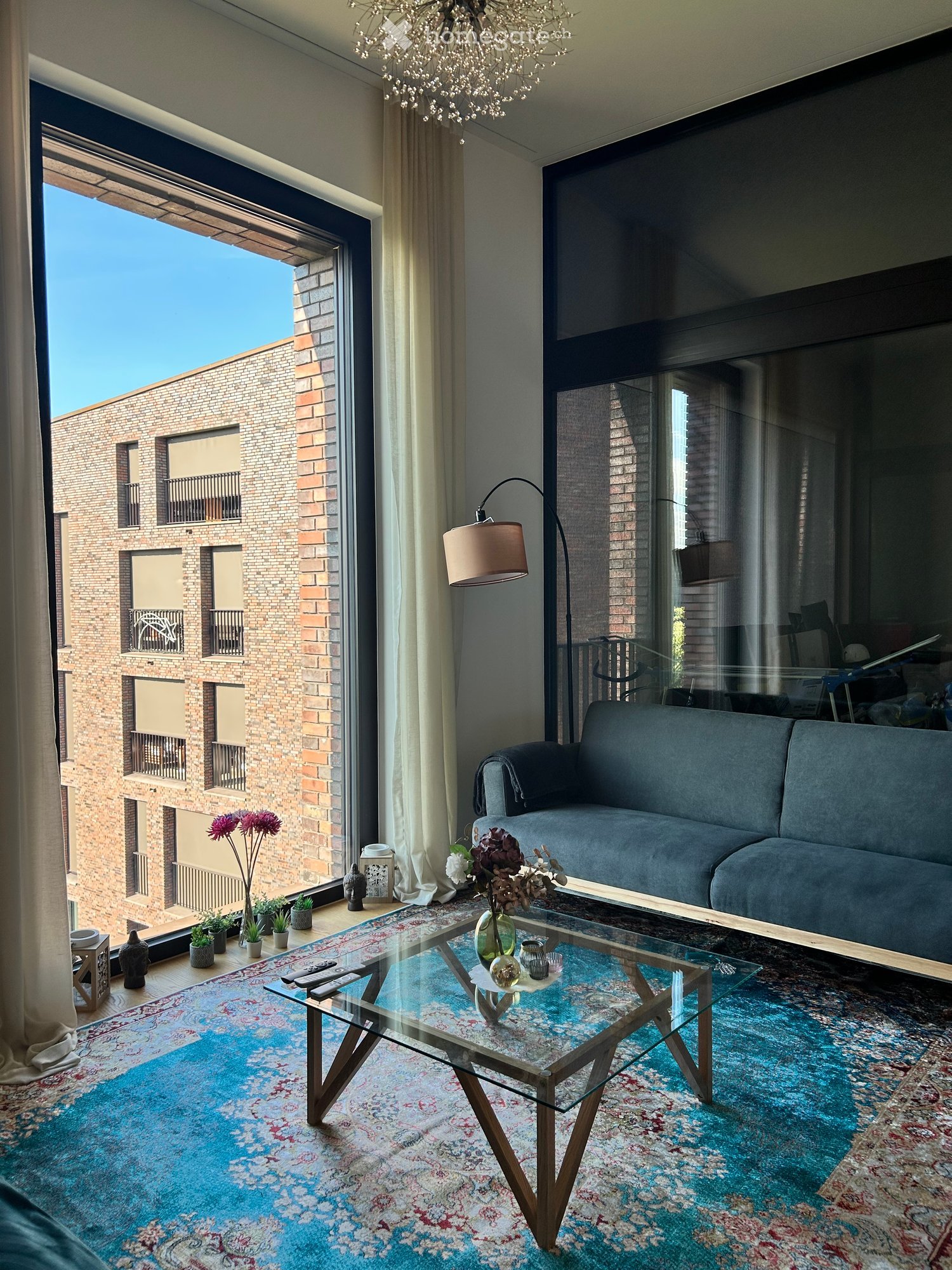 Modern living room with large windows offering a view, a grey sofa, a glass coffee table, a floor lamp, and decorative vases.