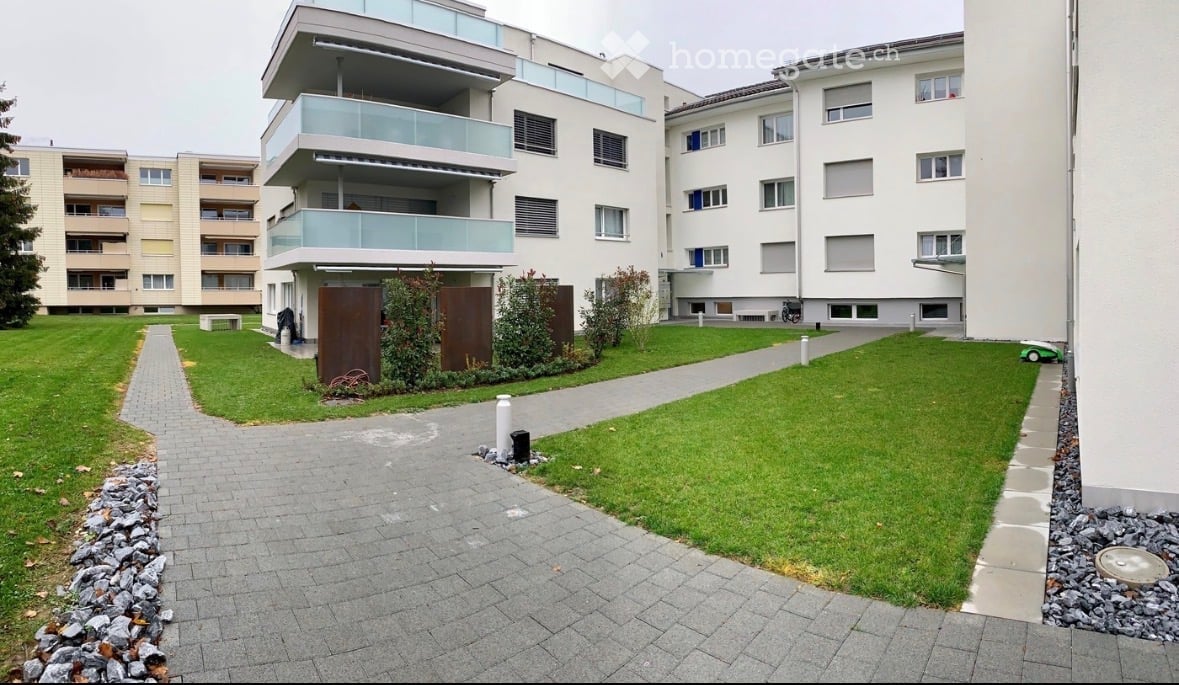 Modern multi-story residential building with balconies, surrounded by a well-maintained lawn and paved pathways.