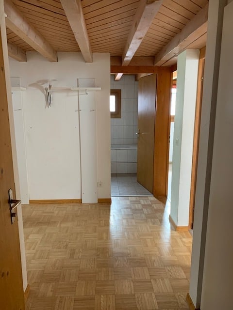 Empty hallway with wooden flooring and white walls, leading to a bathroom with tiled walls and a window.