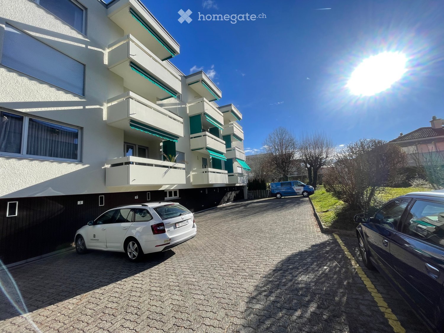White apartment building with green trim, multiple balconies, two parked cars, brick driveway, greenery, and a sunny sky.