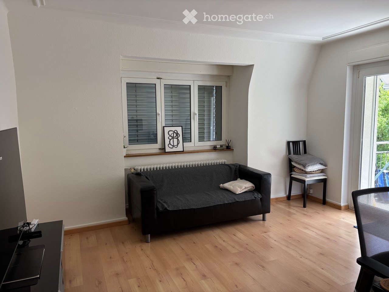 A modern living room with wooden flooring, a black sofa, a small table, a chair, and a picture frame on the wall.