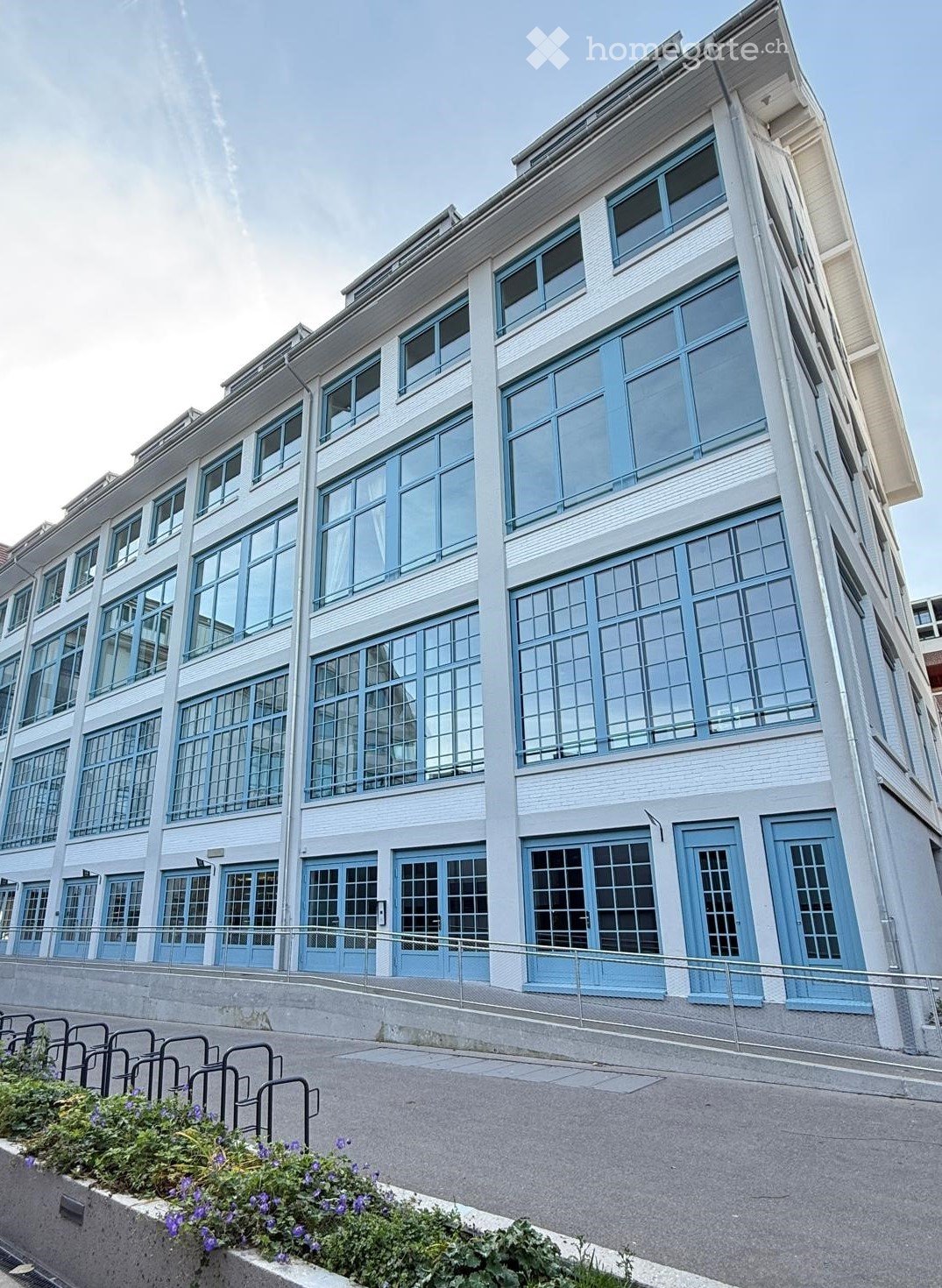 Modern multi-story building with large windows and blue-framed doors, featuring a raised ground floor and a metal railing along the sidewalk.