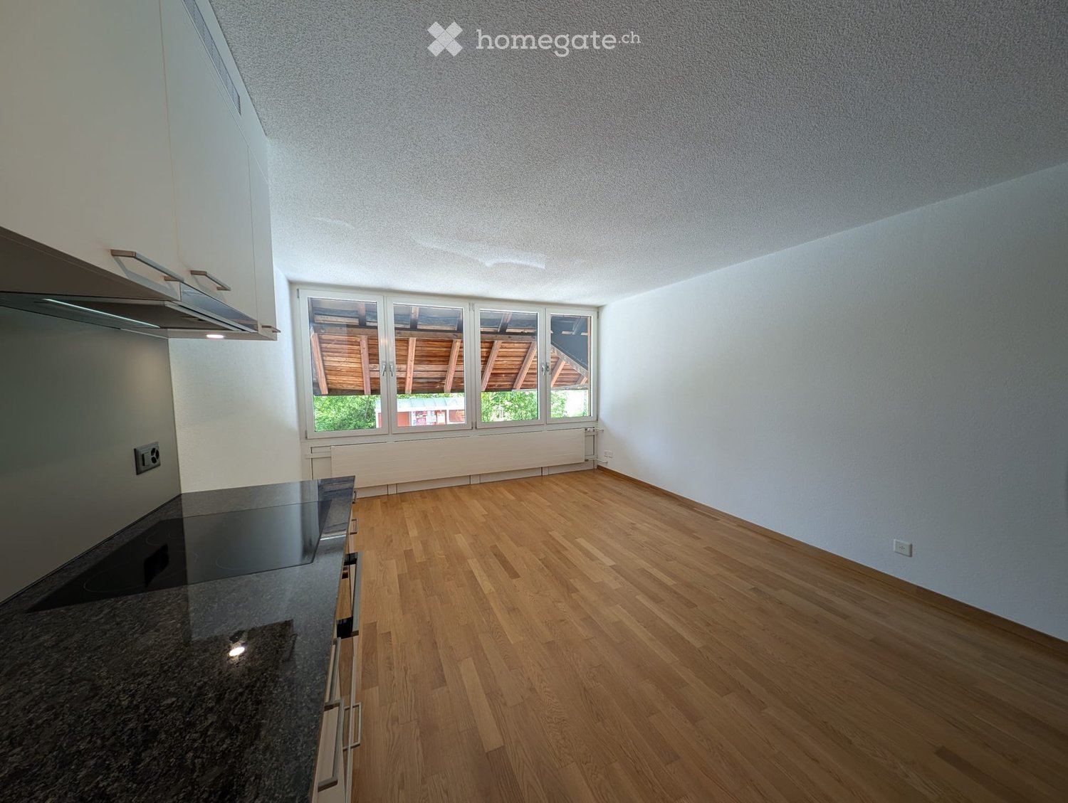 Empty room with wooden flooring, white walls, and a large window with a view of a wooden structure. A modern kitchen counter is visible on the left side.