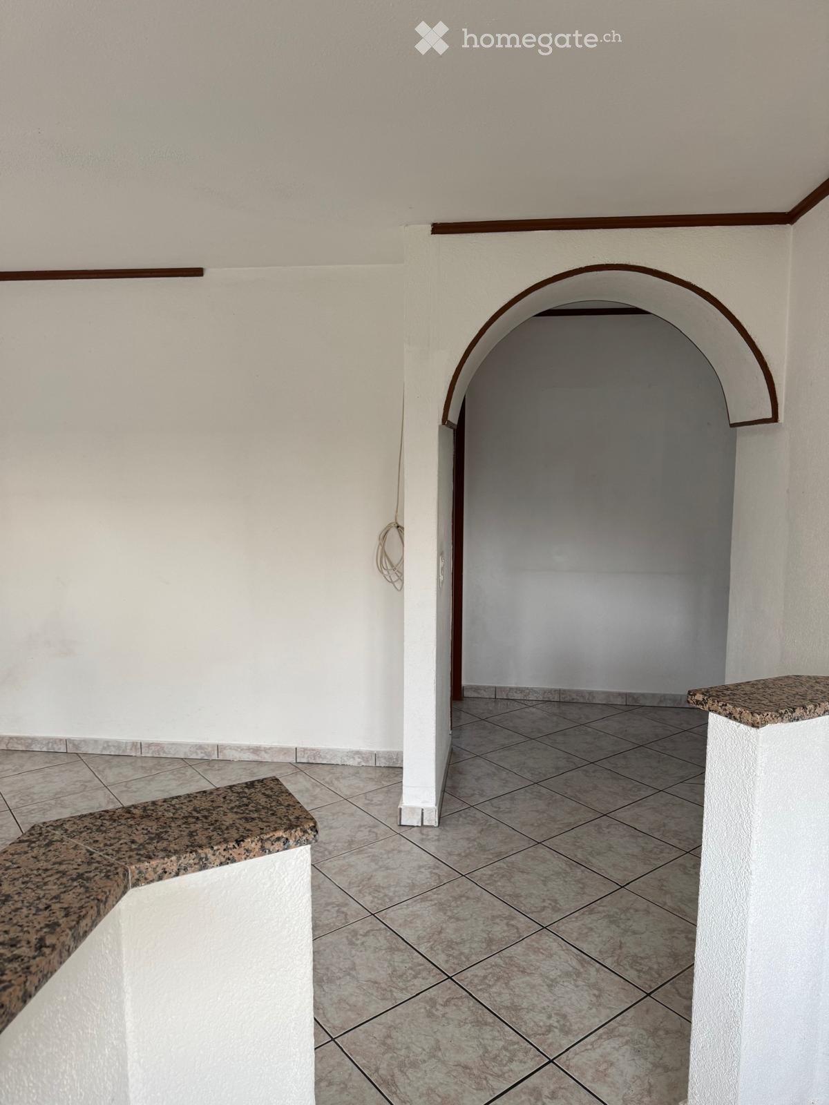 Empty hallway with tiled floor, white walls, and an arched doorway leading to another room. Granite benches on either side.