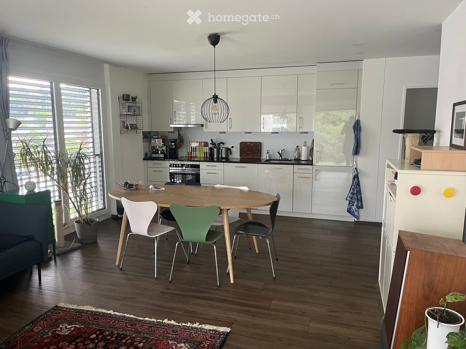 A modern kitchen and dining area with white cabinets, a round wooden dining table, colorful chairs, and various appliances.