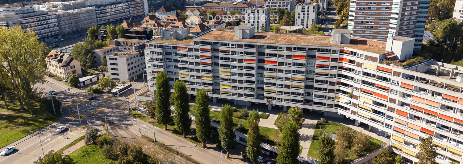 Aerial view of a multi-story residential building with a modern design, featuring a mix of gray and orange accents, surrounded by trees and urban infrastructure.
