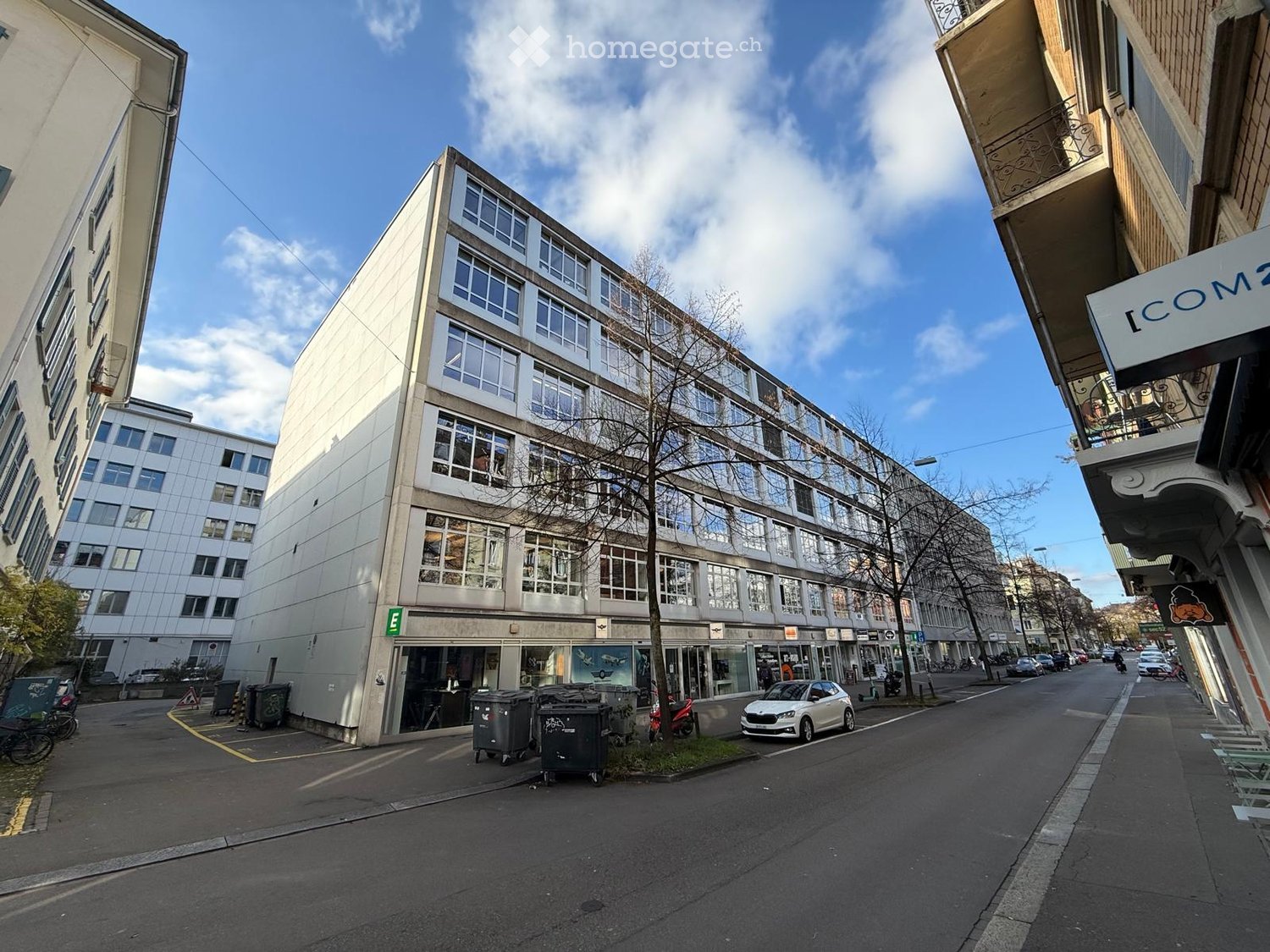 Multi-story building with large windows and a modern facade, located on a street with parked cars and trash bins.