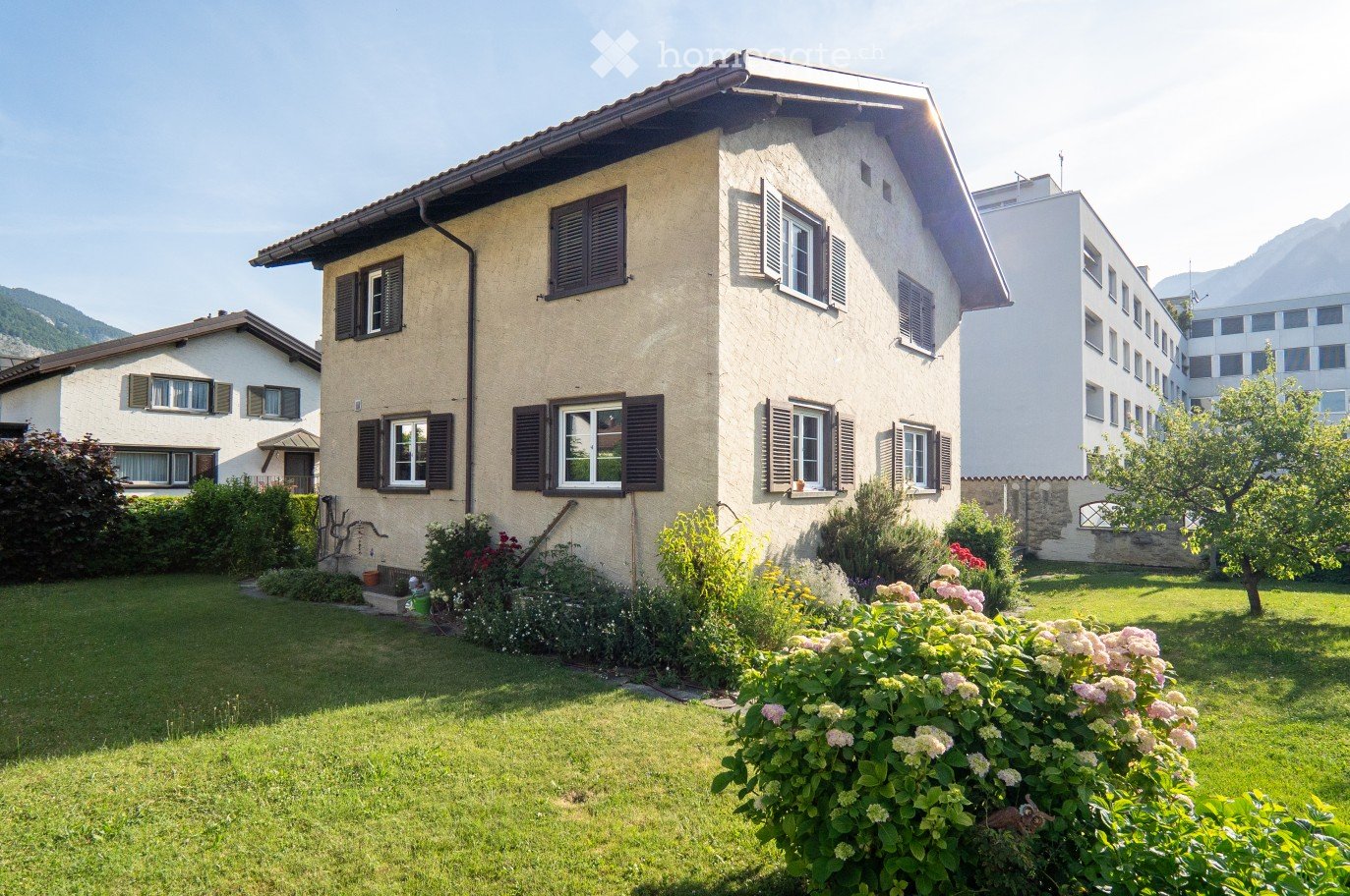 A two-story house with beige walls, dark shutters, and multiple windows. The house is surrounded by a well-maintained garden with green grass, flowering bushes, and trees. The background shows a scenic mountain view.