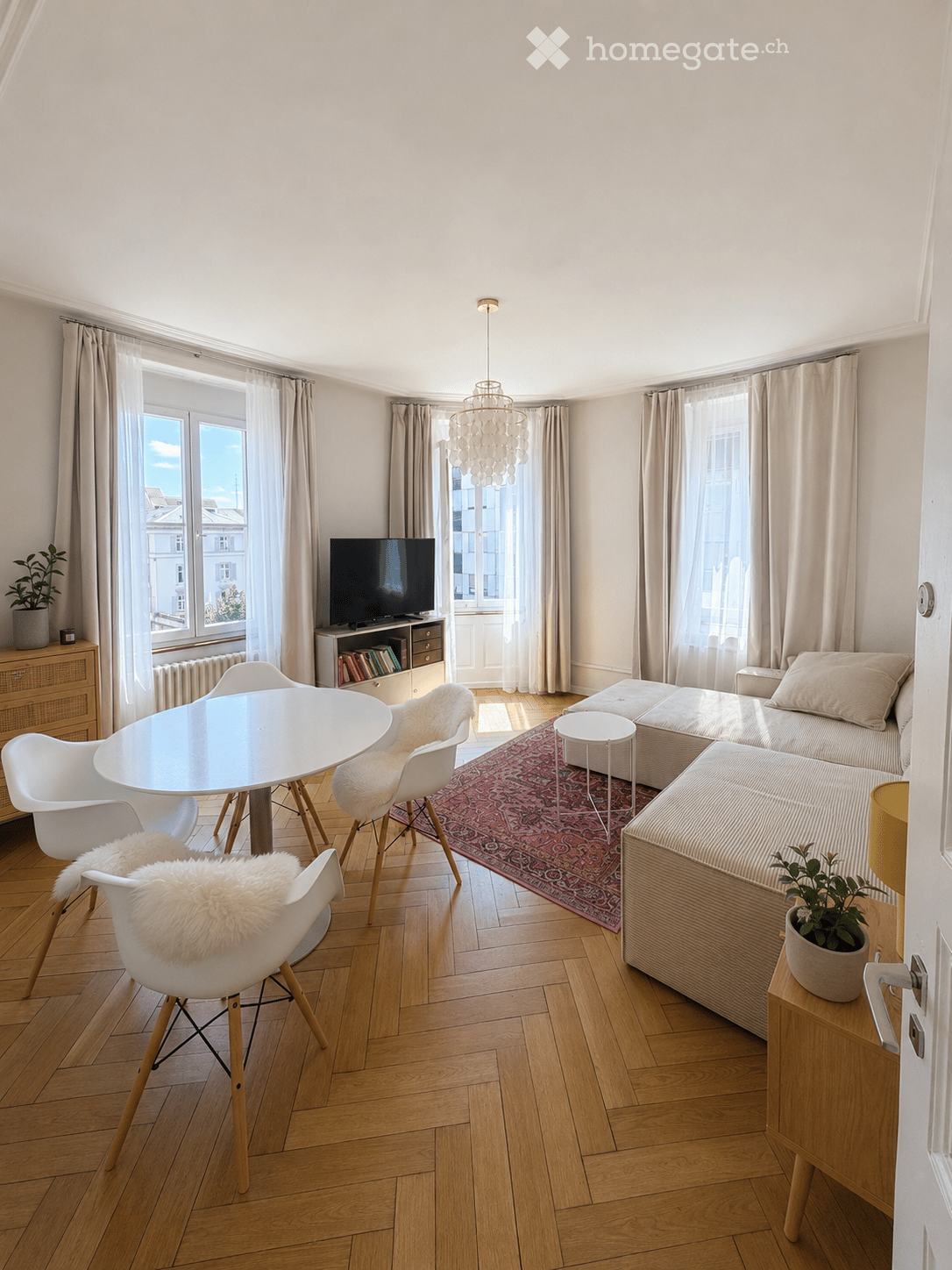 Bright living room with wooden flooring, large windows, a sofa with an ottoman, a coffee table, dining chairs around a table, a TV on a stand, and decorative plants.