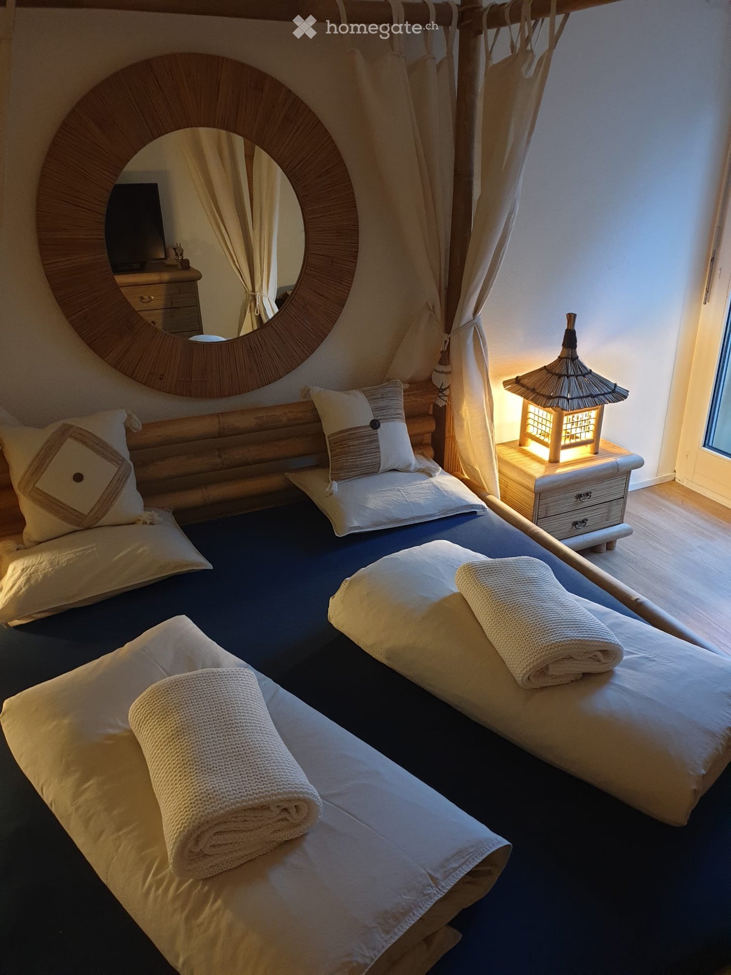 A serene bedroom with a wooden bed frame, neatly arranged pillows and towels, a nightstand with a decorative lamp, and a large round mirror with a wooden frame.