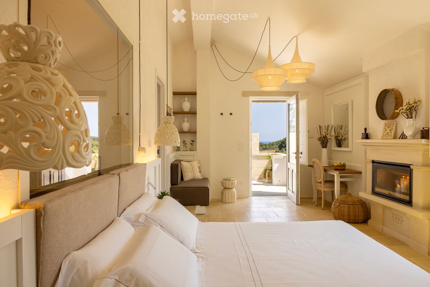 Elegant bedroom with a large bed, white linens, a cozy sofa, a fireplace, and a view of the sea through an open doorway.