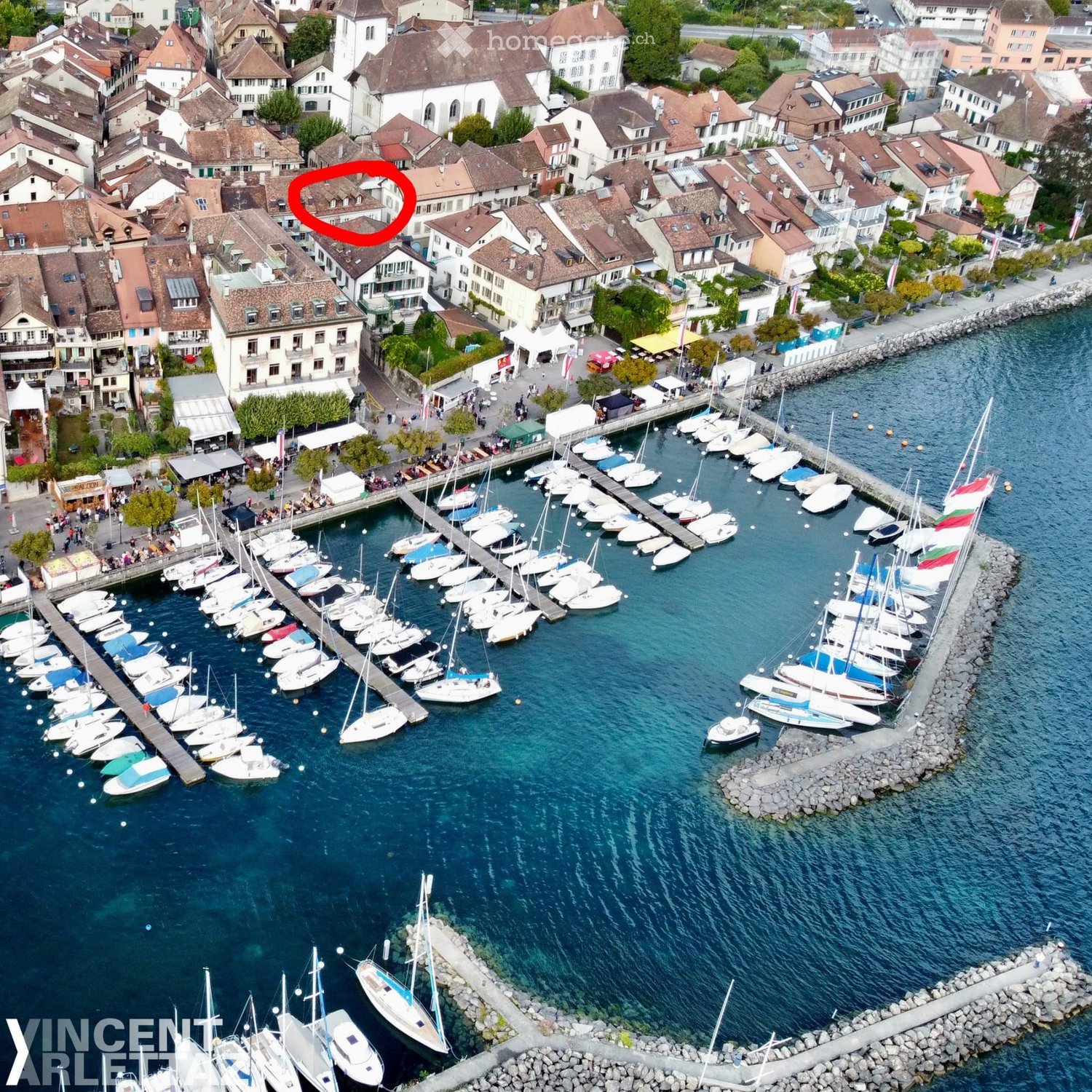Aerial view of a town with a marina filled with sailboats and yachts, surrounded by residential buildings and a waterfront promenade.
