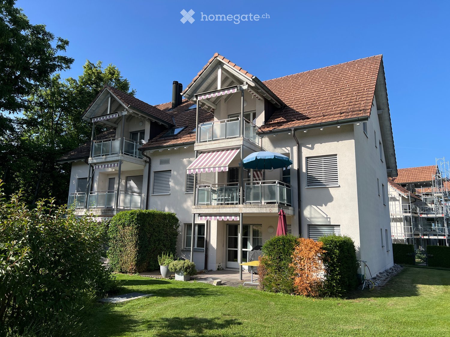 Two-story house with red-tiled roof, white walls, multiple balconies, and a well-maintained garden with lawn, shrubs, and potted plants.