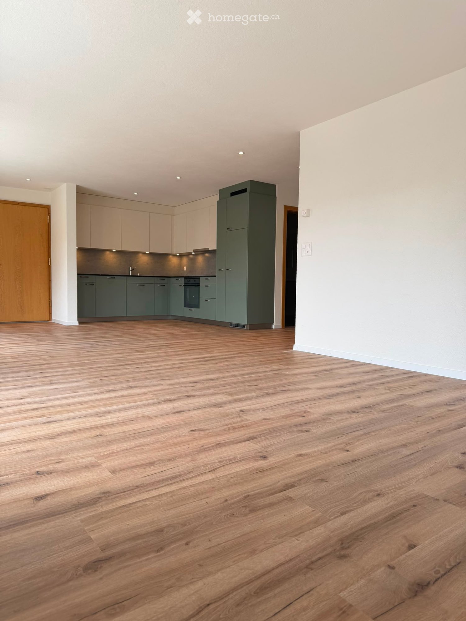 Empty room with wooden flooring, white walls, and a kitchen area visible in the background with cabinets and appliances.