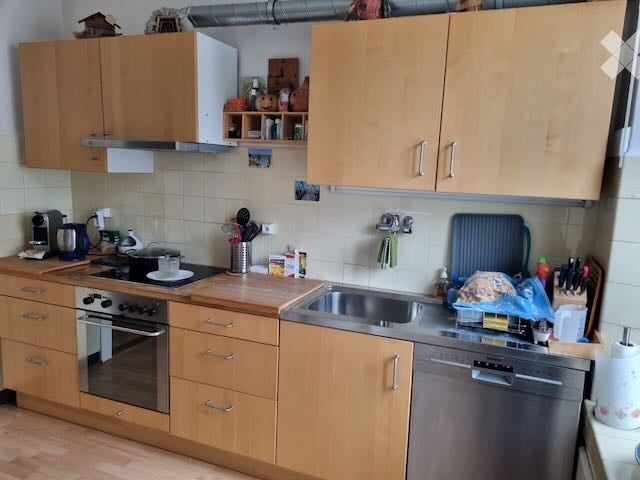 A modern kitchen with light wooden cabinets, stainless steel appliances, and a clean, organized layout. The space features an oven, sink, dishwasher, coffee maker, and various kitchen utensils on the counter.