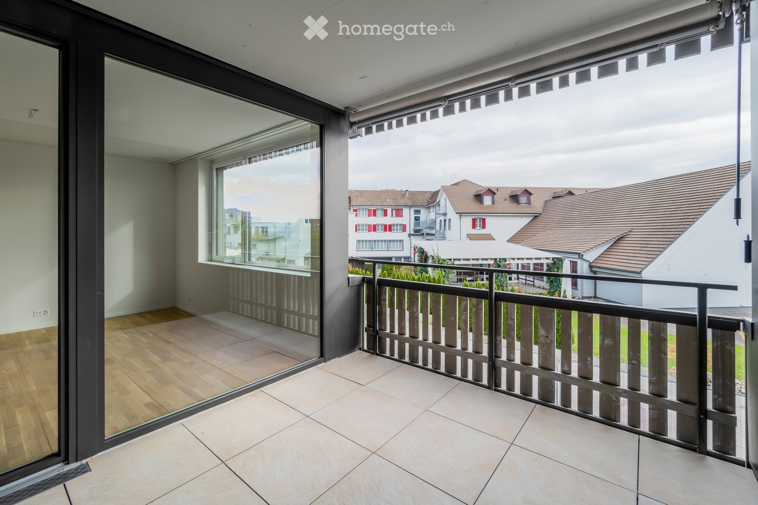 Balcony with tiled floor, wooden railing, large glass sliding doors leading to a room with wooden flooring, and a view of neighboring houses.