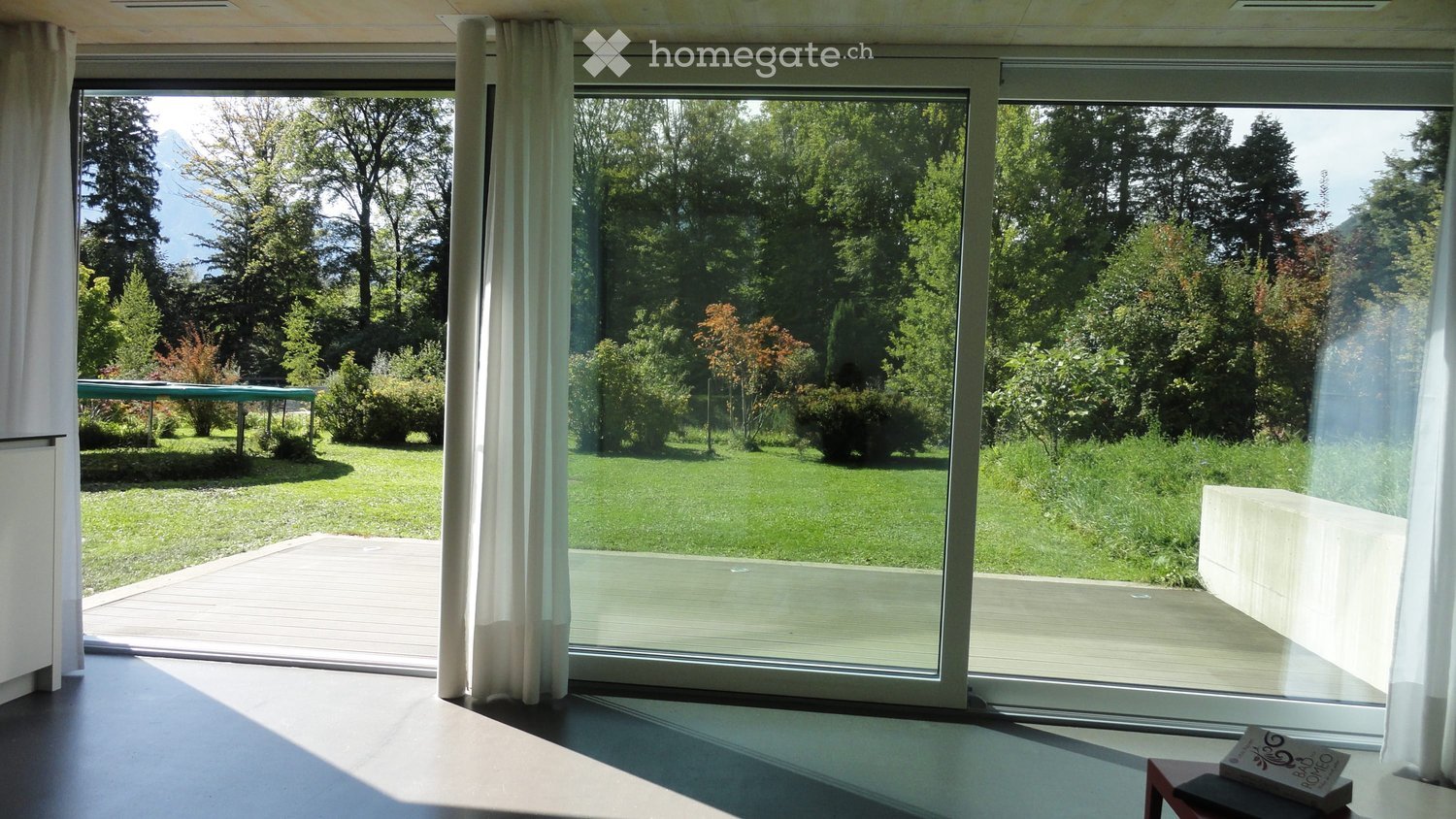 Bright room with large glass sliding doors opening to a green garden, white curtains, a small wooden stool with a book on it, and a small table with a green surface visible through the glass.