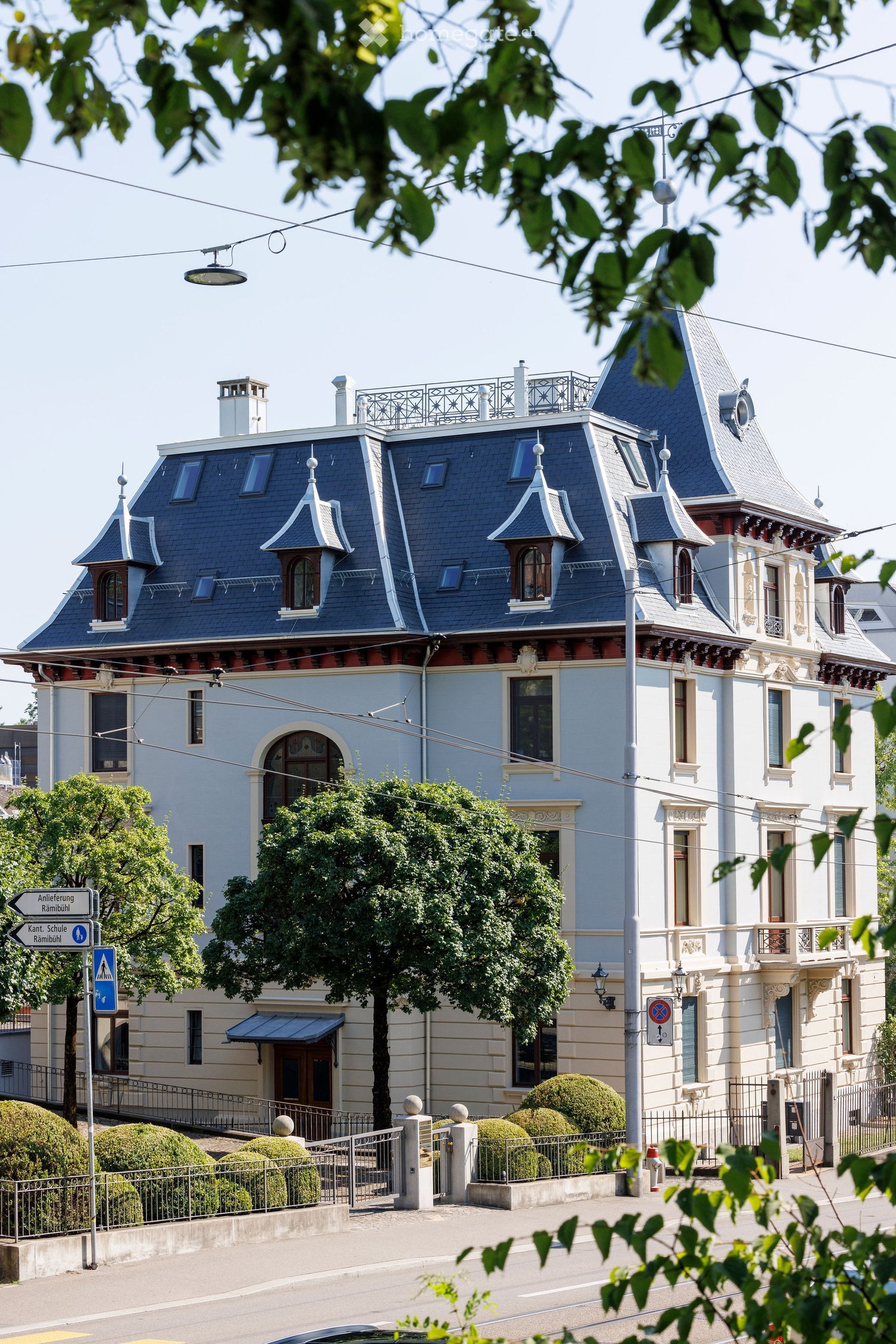 A large, multi-story residential building with a distinctive blue roof and ornate architectural details, surrounded by greenery and a street with traffic signs.