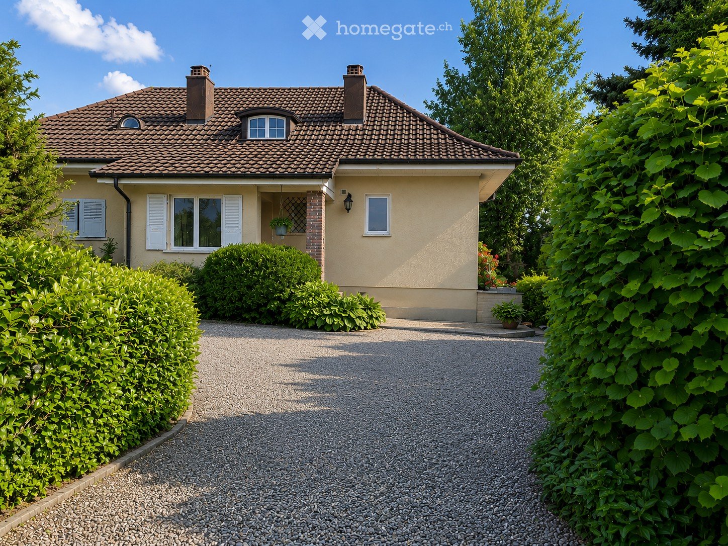 Two-story house with a tiled roof, beige exterior, white shutters, and a gravel driveway surrounded by greenery.