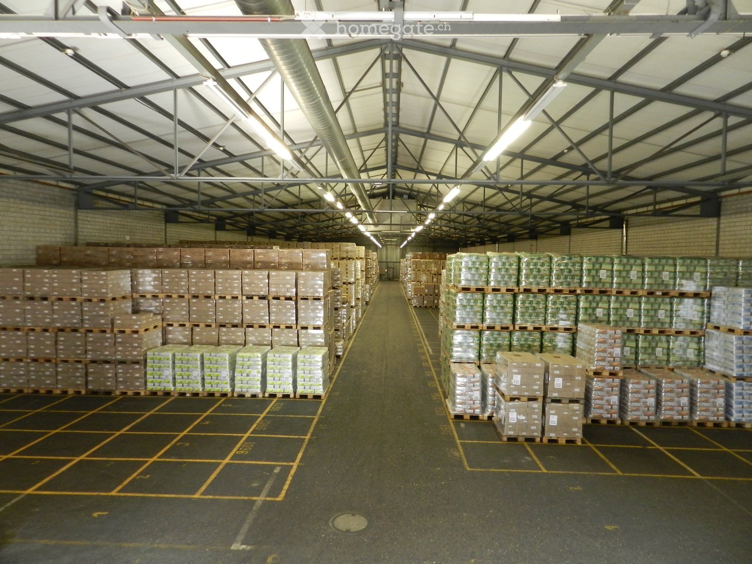 Large industrial warehouse with high ceilings, metal beams, and rows of stacked pallets and boxes on the floor.