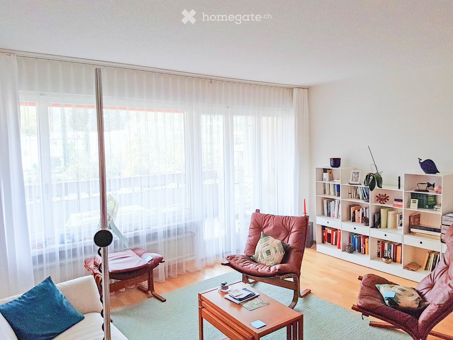 white walls, wooden floor, white curtains, large windows, balcony, armchairs, coffee table, bookshelves, books, plants
