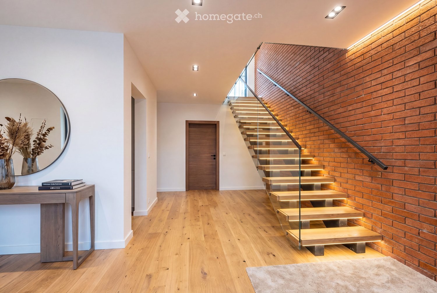 Modern hallway with wooden flooring, a glass staircase with wooden steps, a brick wall, a small table with books and a vase, and a round mirror on the wall.