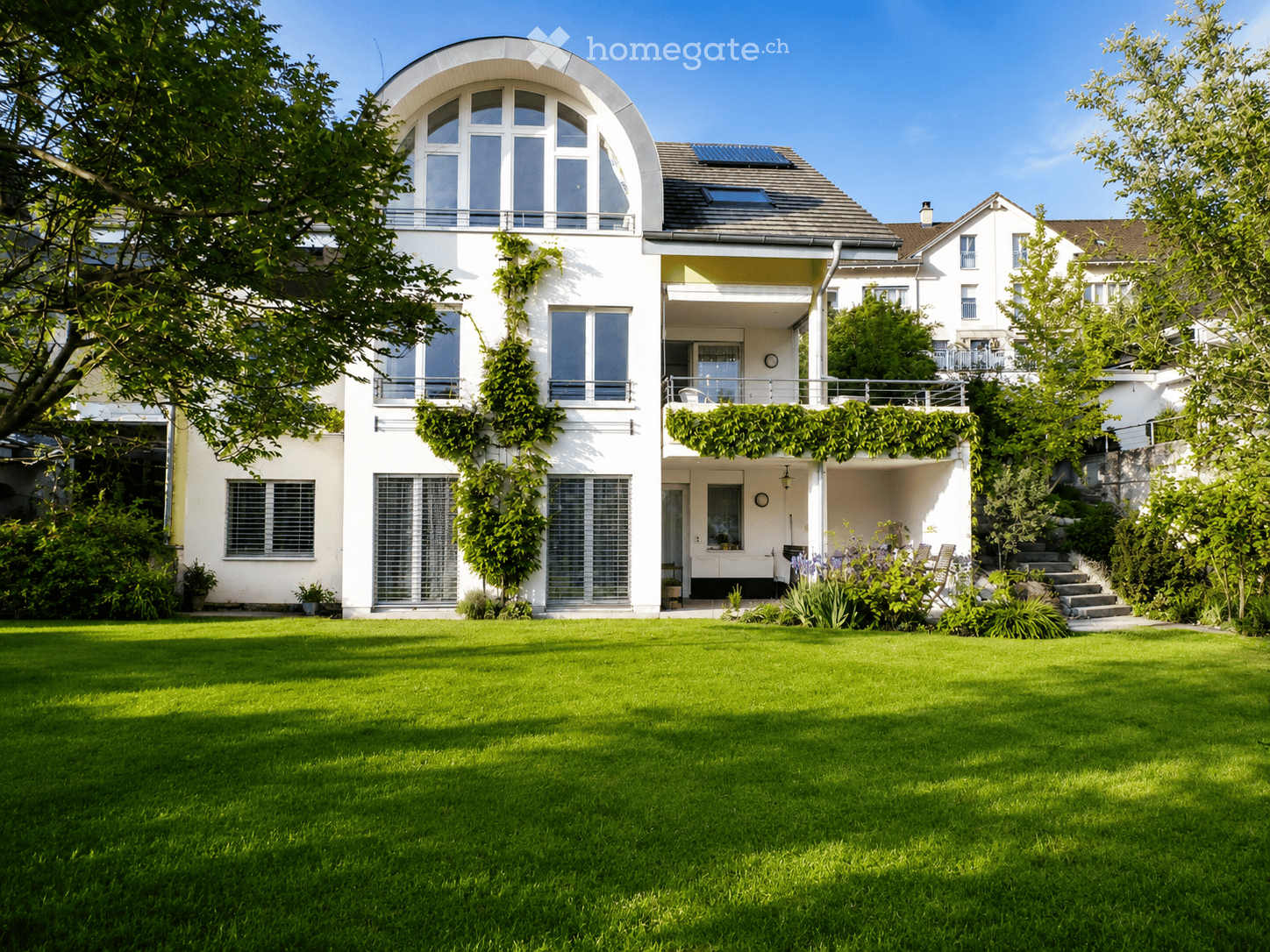 Modern two-story house with white exterior, large windows, and a well-maintained lawn in front. Features include a balcony with greenery, solar panels on the roof, and surrounding trees.