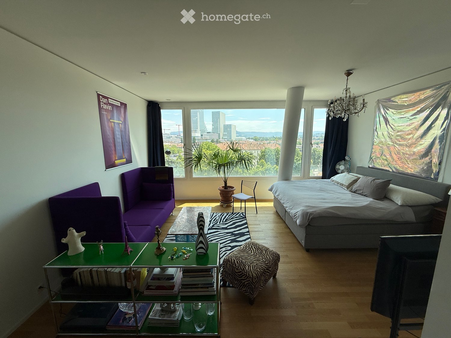 Modern bedroom with a large window offering a city view, featuring a grey bed, purple sofa, glass table with decorative items, bookshelf, and a potted plant.