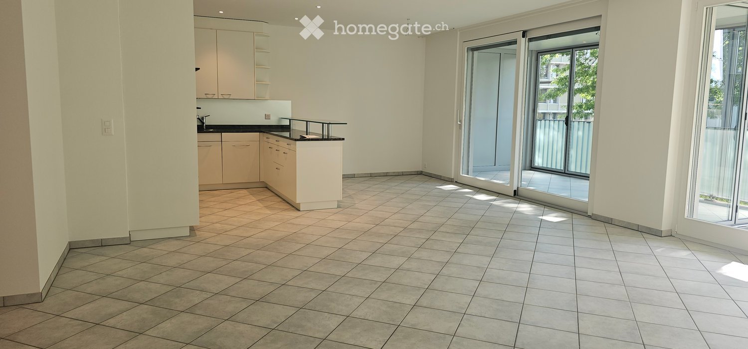 Empty room with tiled flooring, white walls, and a small kitchen area with cabinets and a countertop. Large sliding glass doors lead to a balcony.