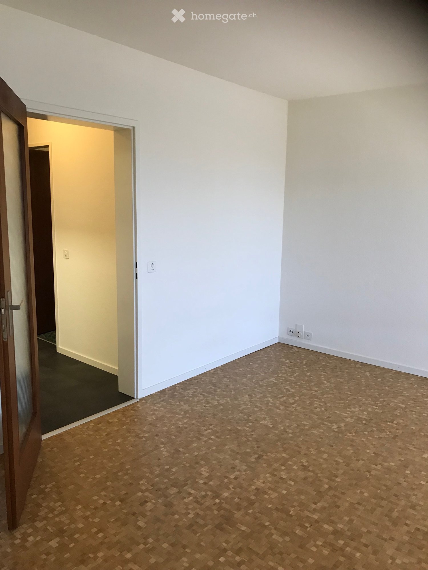Empty room with light-colored walls and a wooden parquet floor. A door leads to another room with a dark floor.