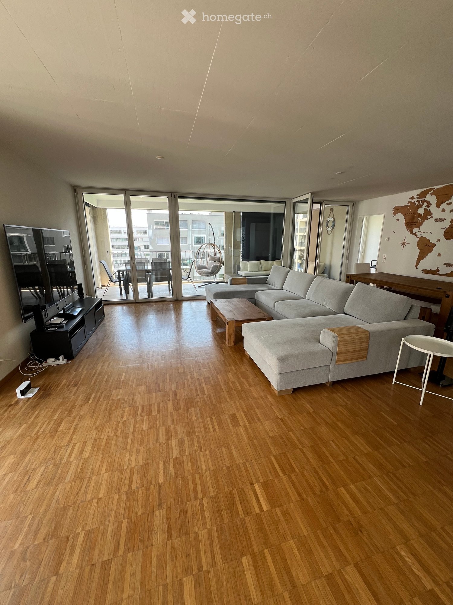 Modern living room with wooden flooring, large sliding glass doors leading to a balcony, grey sectional sofa, coffee table, small round stool, dining table with chairs, TV on stand, and a decorative world map on the wall.