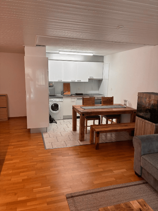 Open-plan living and dining area with a sofa, dining table, bench, chairs, and a small kitchen with a washing machine, fridge, and oven.