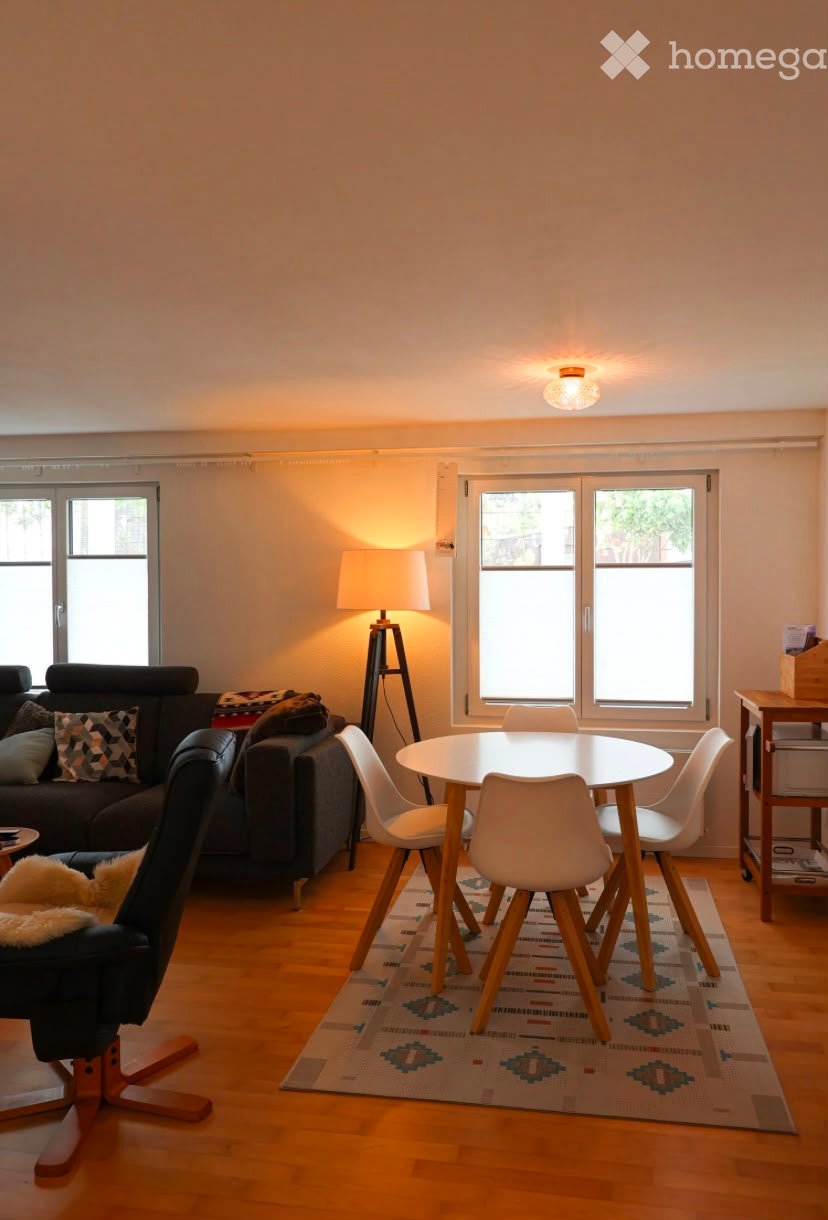 Bright living room with wooden flooring, large windows, a sofa, a round dining table with chairs, a floor lamp, and an ottoman.