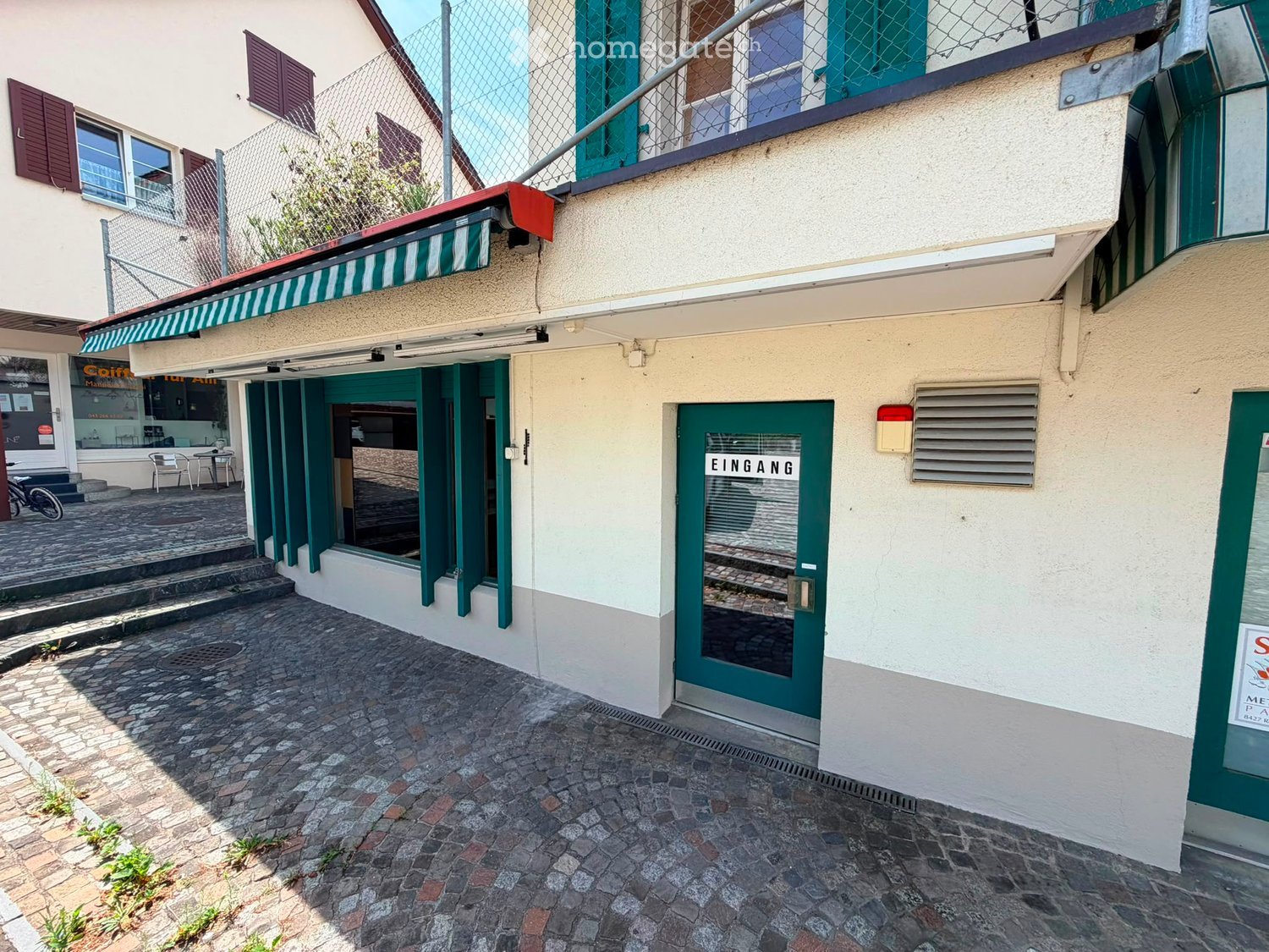 Two-story building with green shutters, a green entrance door labeled "EINGANG", and a small awning over a storefront. The ground floor has a paved stone walkway and a small outdoor seating area with tables and chairs.