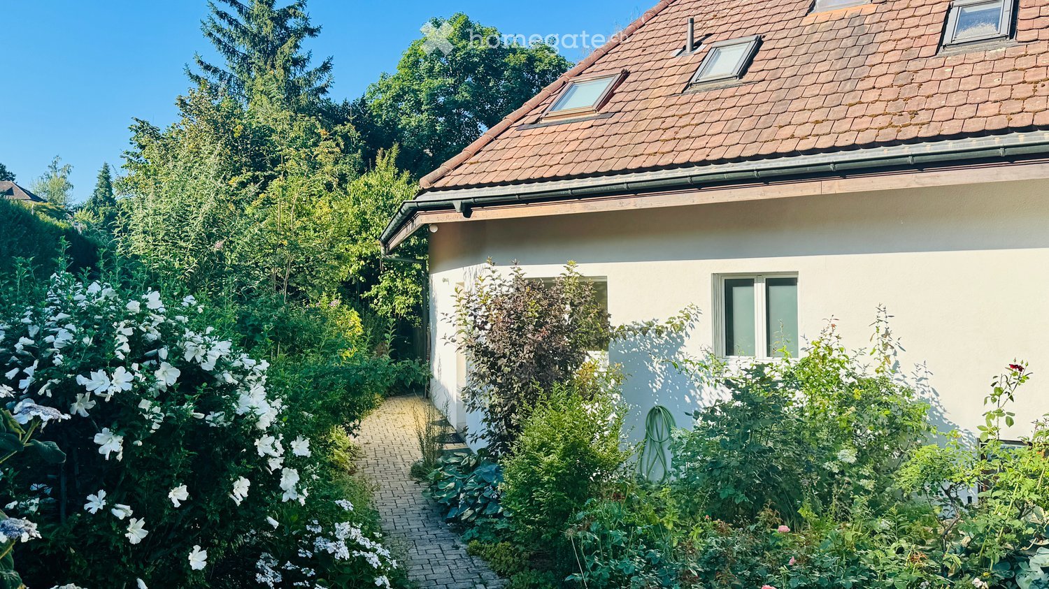 A well-maintained garden with a pathway leading to a white house with a red-tiled roof. The garden features lush greenery, flowering shrubs, and a neatly trimmed hedge.