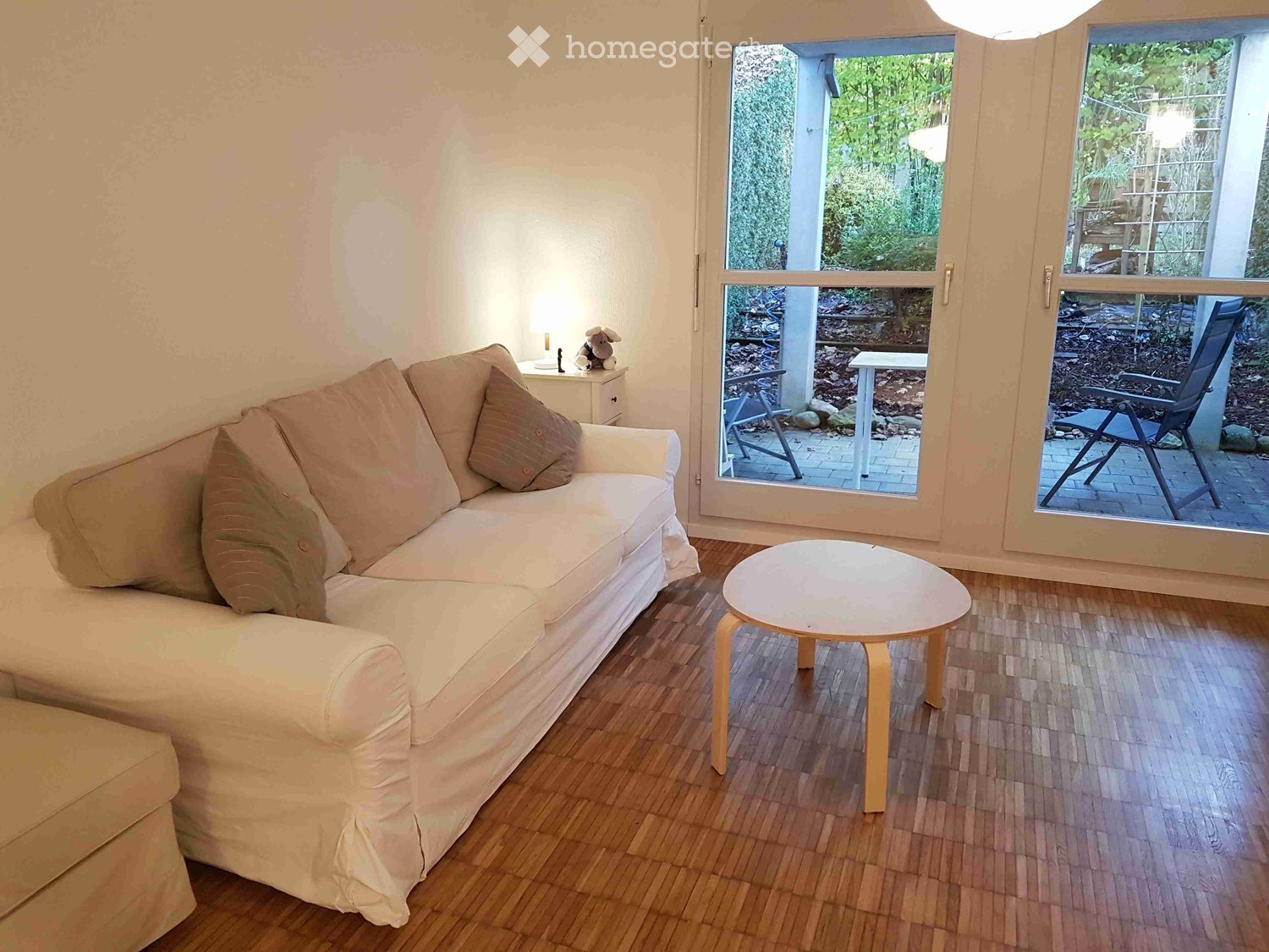 Living room with a white sofa, coffee table, side table, and a small stool. Large sliding glass doors open to a patio with outdoor furniture.