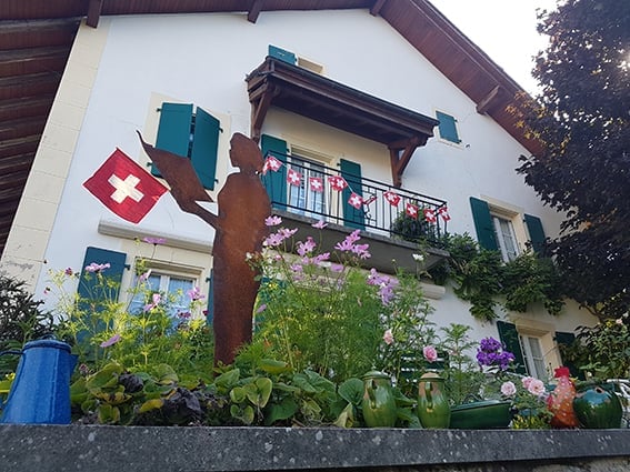 A charming white house with green shutters, a balcony adorned with flowers and a Swiss flag, and a decorative metal sculpture in the foreground.