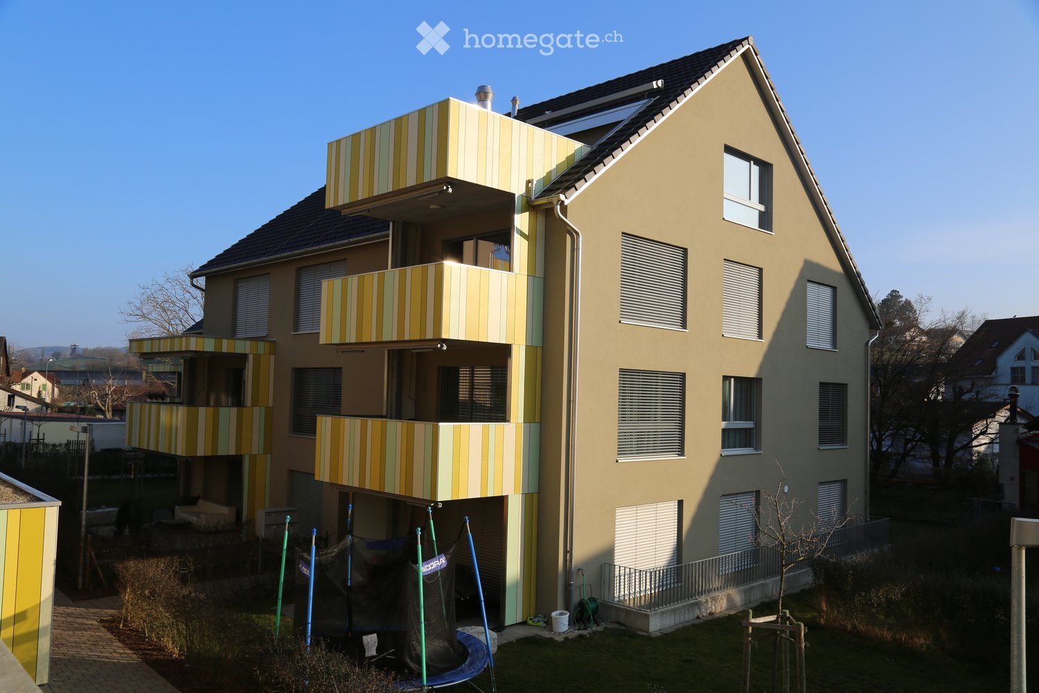 Modern multi-story residential building with yellow and green striped balconies, multiple windows, and a flat roof. The building is surrounded by a small garden area with a trampoline and some greenery.