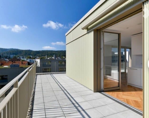 Balcony with tiled floor, metal railing, and sliding glass door leading to a room with wooden flooring and white cabinets.