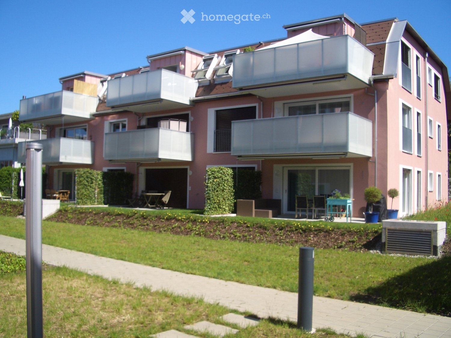 Pink multi-story residential building with multiple balconies, modern design, and well-maintained garden area with outdoor furniture.