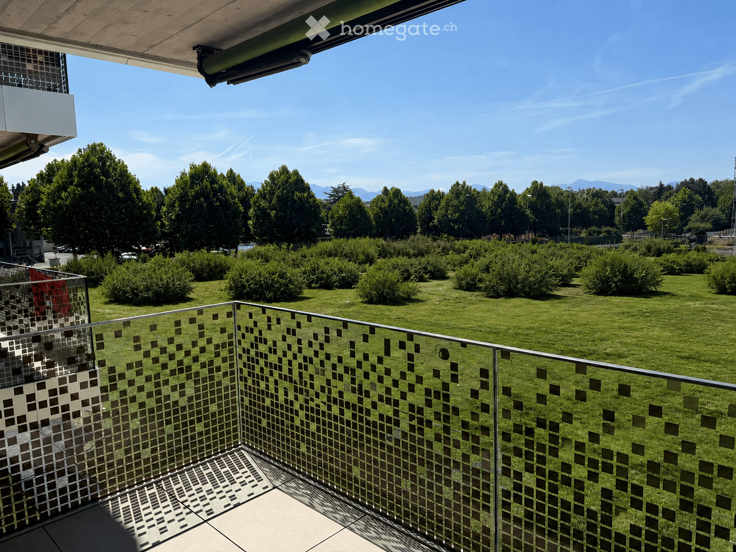 Balcony with modern glass railing overlooking a green lawn and trees, with a clear blue sky and distant mountains in the background.