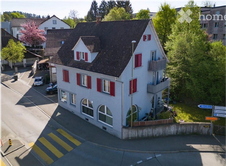 A two-story house with a dark roof and red shutters, surrounded by greenery and parked cars.