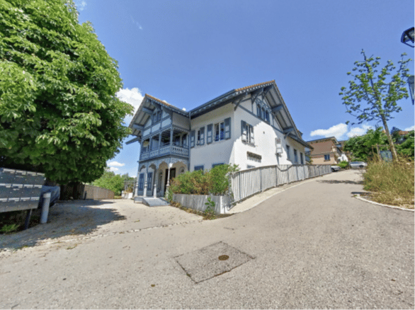 A two-story white house with blue trim and decorative wooden elements, situated on a raised ground floor with a gravel driveway and small garden area.