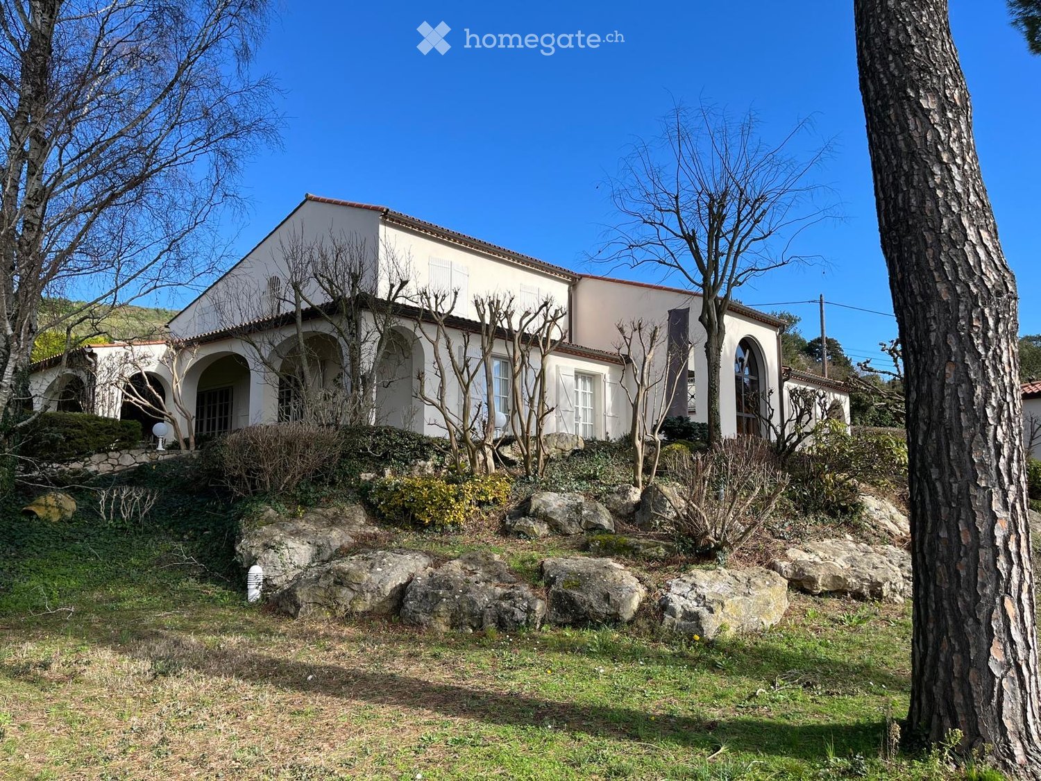 A two-story white house with arched entrances and windows, surrounded by a landscaped garden with rocks and trees.