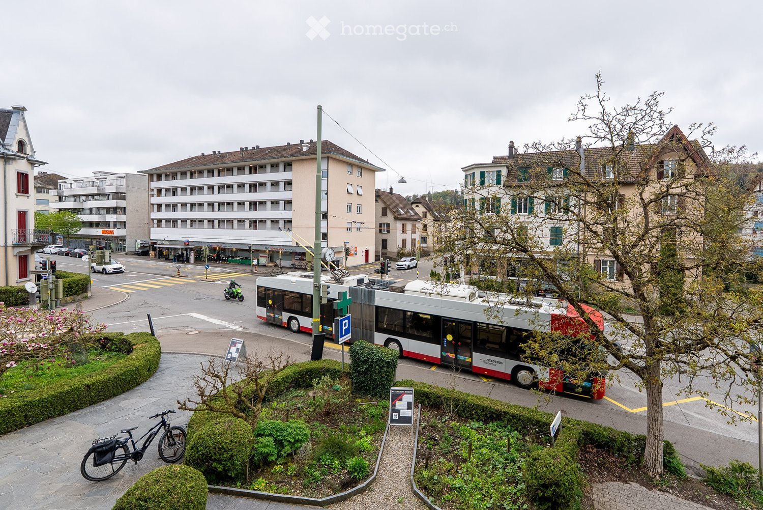 Urban street scene with multi-story buildings, a bus, cars, and a bicycle in a landscaped area.