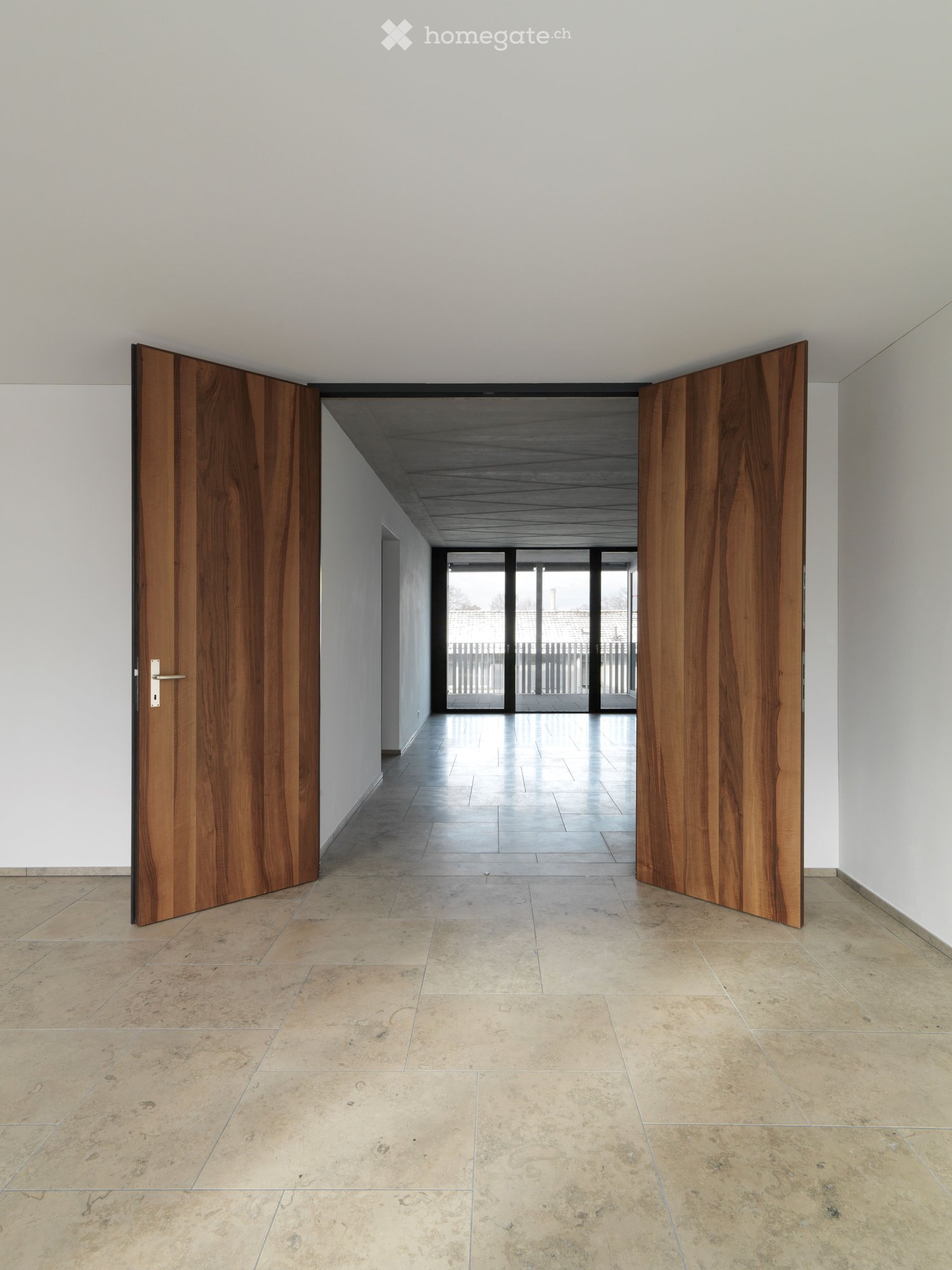 Empty hallway with light-colored tiled flooring, white walls, and large wooden double doors leading to an outdoor area with glass panels.