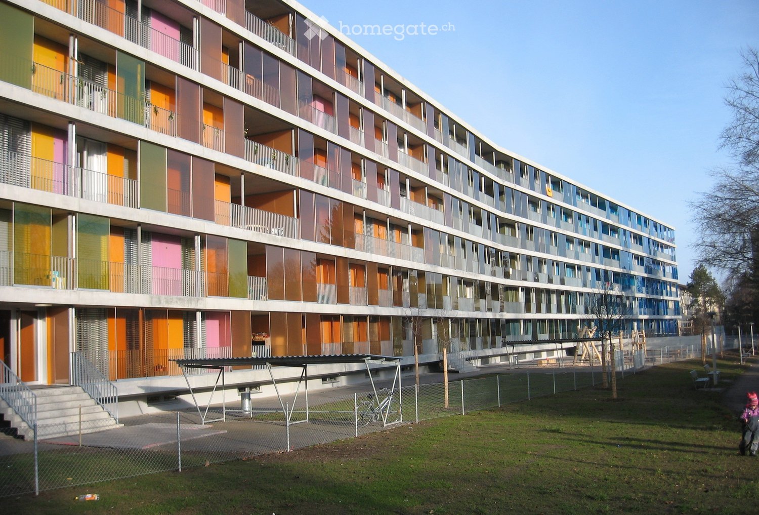 A modern multi-story residential building with colorful panels and large windows, featuring balconies and a fenced playground area in front.