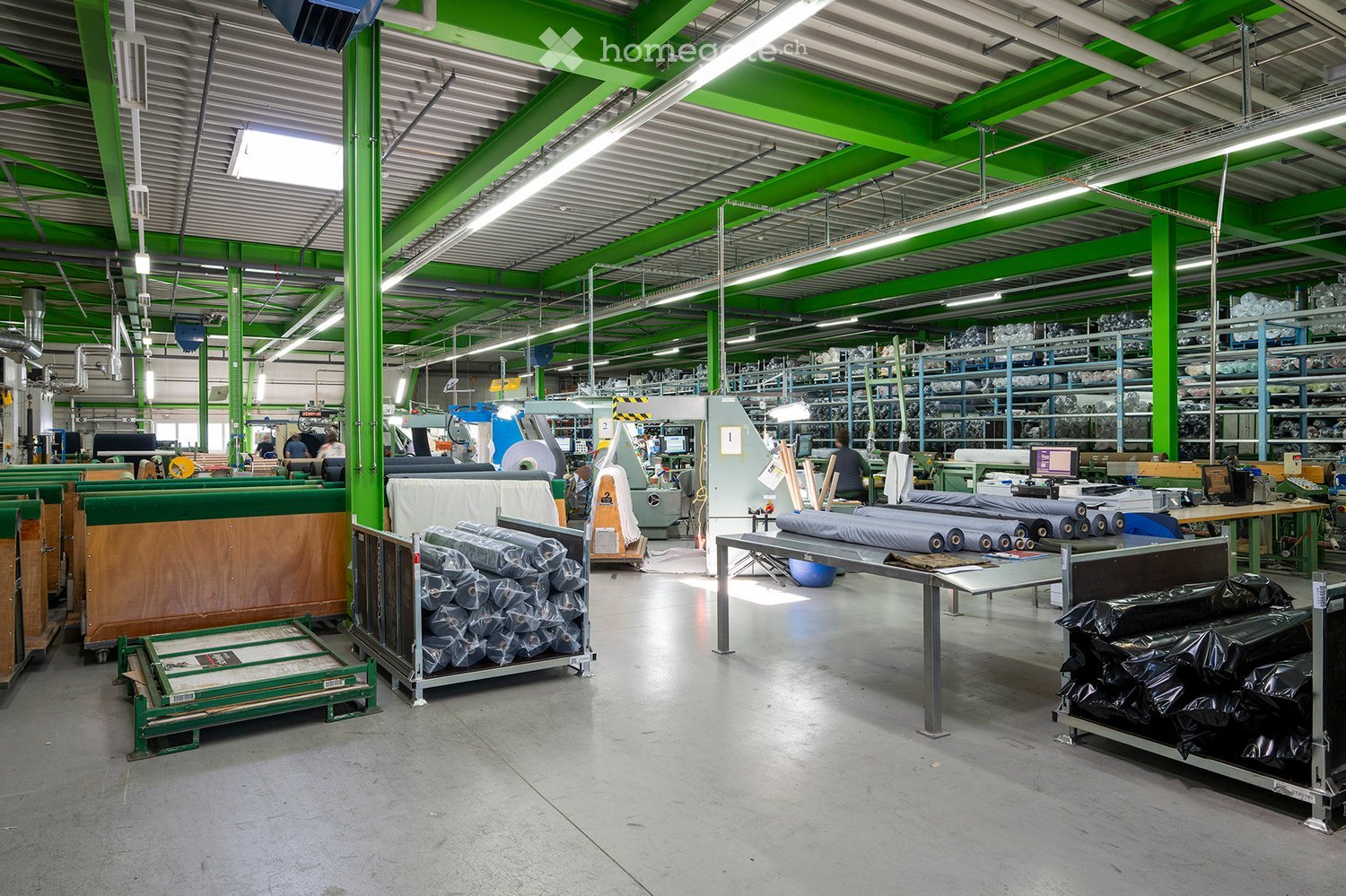 Large industrial warehouse with green structural beams, metal shelving units, and various machinery. The space features a high ceiling with exposed lighting and ventilation ducts, with rolls of material and equipment visible throughout.
